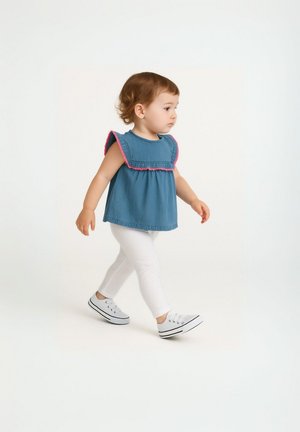Tout-petit avec des cheveux bouclés marchant, portant un haut sans manches bleu avec des bordures roses, des leggings blancs et des baskets blanches sur un fond uni.