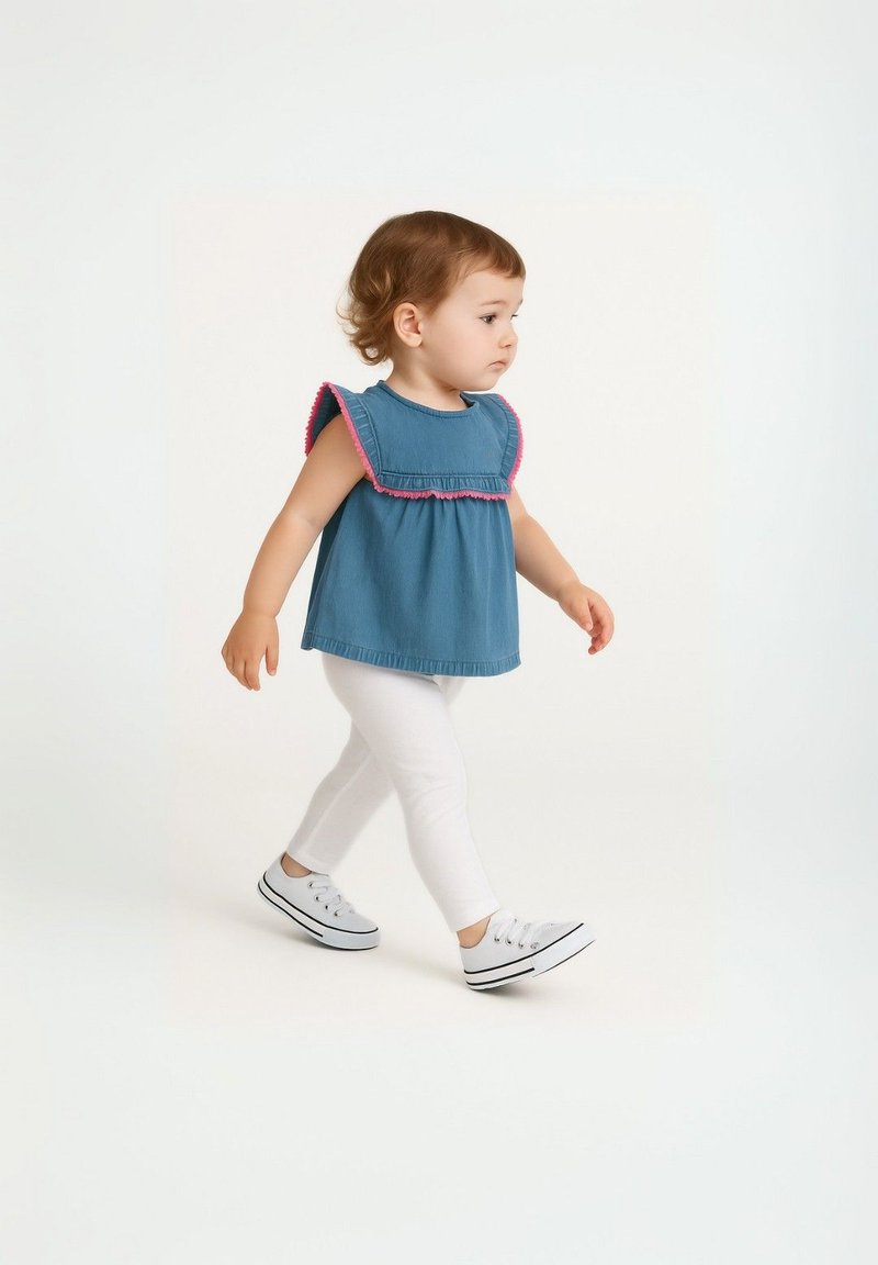 Bambino con capelli ricci che cammina, indossando una maglietta senza maniche blu con bordi rosa, leggings bianchi e sneaker bianchi su uno sfondo neutro.