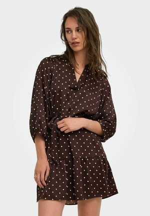 Femme aux cheveux bruns lâchés portant une robe marron à pois avec des manches trois-quarts et une ceinture nouée à la taille, regardant sur le côté.