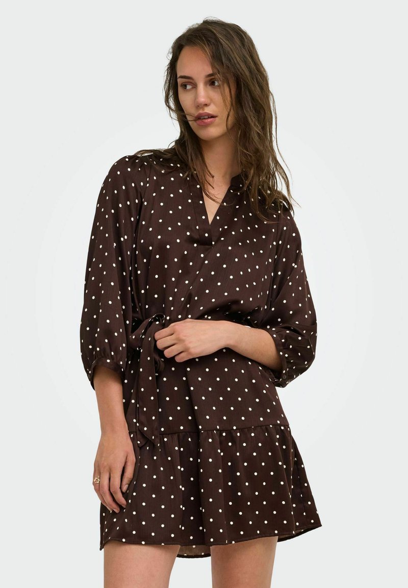 Femme aux cheveux bruns lâchés portant une robe marron à pois avec des manches trois-quarts et une ceinture nouée à la taille, regardant sur le côté.