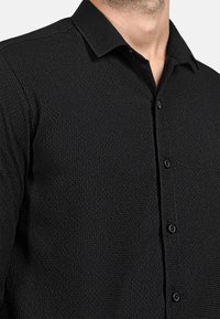 Camicia nera a maniche lunghe con colletto, caratterizzata da un motivo a tessitura compatta e una lieve lucentezza, con bottoni in plastica nera.