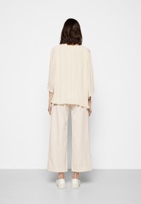 Beige, gefältete Bluse mit langen Ärmeln, kombiniert mit weiten, beigen Hosen. Das Outfit hat eine lockere Passform und einen weichen, strukturierten Stoff.