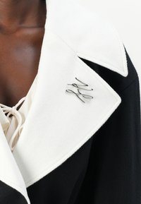 Manteau en laine bicolore noir et blanc avec un large col blanc et un broche argentée au design torsadé. Tissu texturé visible.