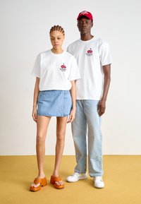 Hvit t-skjorte med kirsebærgrafikk, lys denimskjørt og oransje platåsandaler. Grå jeans og hvite sneakers. Plain bakgrunn.