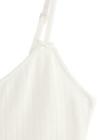 Wit geribd gebreid tanktopje met dunne verstelbare bandjes en een klein decoratief strikje vlak bij de halslijn.