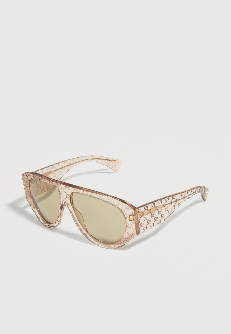 Lunettes de soleil transparentes beige forme œil de chat avec un motif monogramme subtil et des verres teintés marron clair sur un fond blanc.