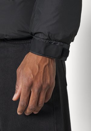 Gros plan d'une main portant une veste noire à manches longues avec un poignet à fermeture réglable en crochet et boucle, sur un pantalon foncé.