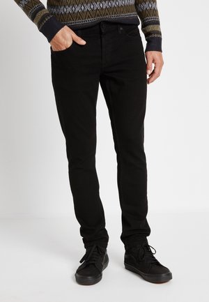 Mand iført sorte slim-fit jeans og sorte sneakers, med den ene hånd i lommen og mønstret sweater synlig fra taljen og ned.
