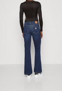 Henkilö, jolla on tummansiniset leveälahkeiset farkut, musta pitkähihainen paita ja valkoiset lenkkitossut mustilla kuvioilla, seisoo yksinkertaista valkoista taustaa vasten.