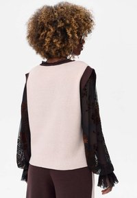 Beżowy top bez rękawów z dzianiny, nałożony na czarną przezroczystą bluzkę z długim rękawem z kwiatowym haftem. Całość dopełniają brązowe textured spodnie.