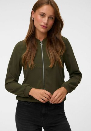 Femme portant une veste verte zippée et un pantalon noir, debout, mains jointes à la taille, sur un fond clair et uni.