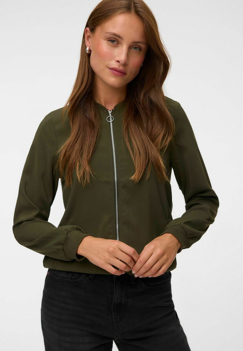 Femme portant une veste verte zippée et un pantalon noir, debout, mains jointes à la taille, sur un fond clair et uni.