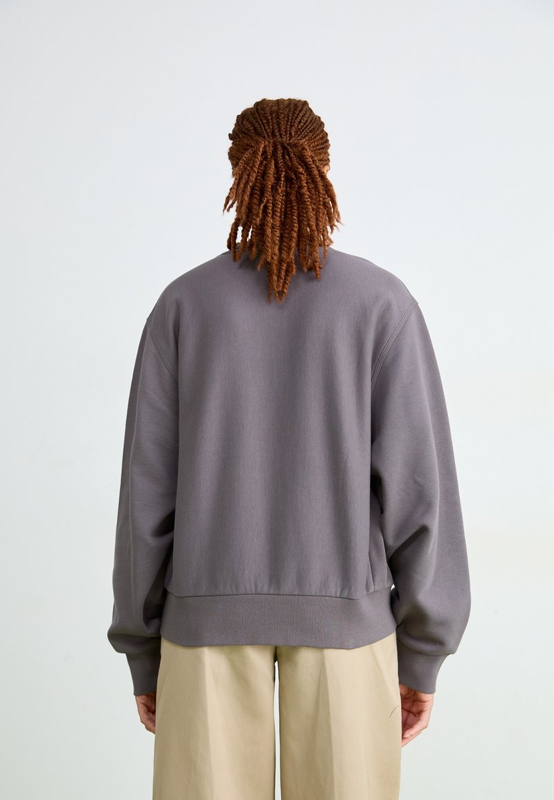 Grå sweatshirt med avslappnad passform, ribbade muddkanter och fåll. Slät textur, rund hals och sänkta axlar. Bärs över ljusa byxor.