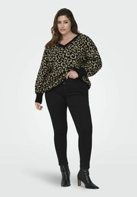 Moteris, vilkinti leopardo rašto megztinį, juodas prigludusias kelnes ir juodus aukštakulnius aulinukus, stovi prieš paprastą foną.