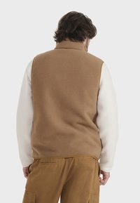 Chaleco de forro polar con espalda marrón y mangas blancas, que presenta un cuello alto y una textura sutil. Se lleva con pantalones color tostado, mostrando un estilo casual.