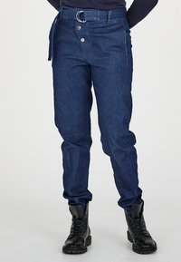 Mörkblå denimbyxor med hög midja, utrustade med ett runt bältesspänne, sidoknappar och en avsmalnande benform. Svarta stövlar kompletterar looken.