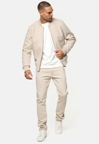 Beige bomber jacket with ribbed cuffs, worn over a white sweatshirt and light beige pants. Completed with white sneakers.