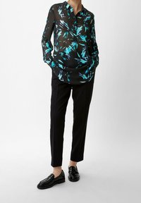 Blouse noire transparente avec un motif abstrait turquoise, manches longues et col, assortie à un pantalon noir ajusté et des mocassins brillants.