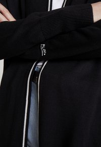 Černý pletený kardigan s bílými akcenty, se zipovým uzávěrem a žebrovanými manžetami. Na levé straně výrazné logo.