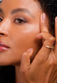 Femme à la peau lisse touchant doucement le côté de son visage près de la tempe avec ses doigts, portant une bague en or à son doigt.