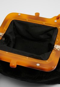 Kabelka s akrylovým rámem v jantarové barvě a měkkým černým látkovým interiérem, s kovovým zapínáním a vyraženým logem značky.