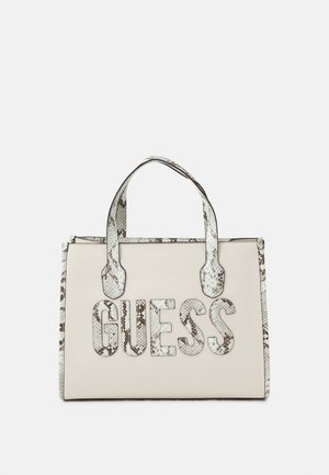Kremowa torba typu tote z ozdobami i uchwytami z węża, z dużym, teksturowanym logo "GUESS" z przodu.