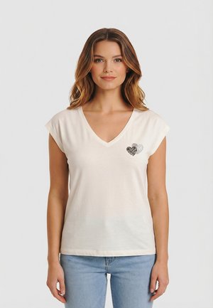 Vrouw draagt een wit V-hals topje met korte kapmouwen en twee kleine hartjes op de borst, gecombineerd met lichtblauwe jeans, staand tegen een effen achtergrond.