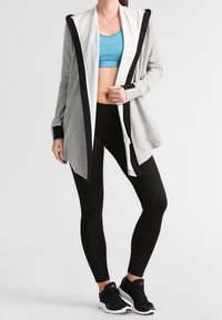 Cárdigan gris con capucha y acentos negros, usado sobre un sujetador deportivo azul y leggings negros a rayas, combinado con zapatillas deportivas negras.