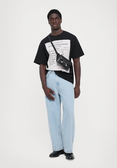 T-shirt graphique noir avec impression, jean droit bleu clair, et sac bandoulière noir. Le modèle se tient sur un fond blanc.