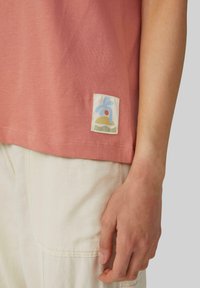 Bawełniana koszulka w delikatnym różu z kwadratową, tkana metką z niebieskim i żółtym wzorem. Noszona z jasnobeżowymi spodniami, ukazującymi strukturę.