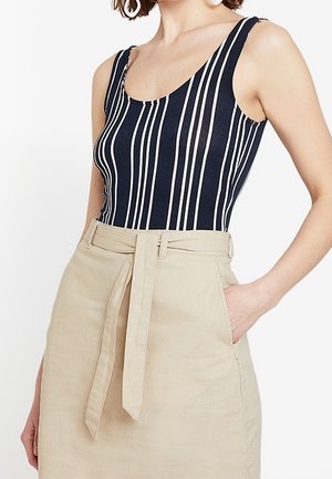 Vrouw die een mouwloze marineblauwe top met verticale witte strepen draagt, ingestopt in een beige rok met riem en zakken.