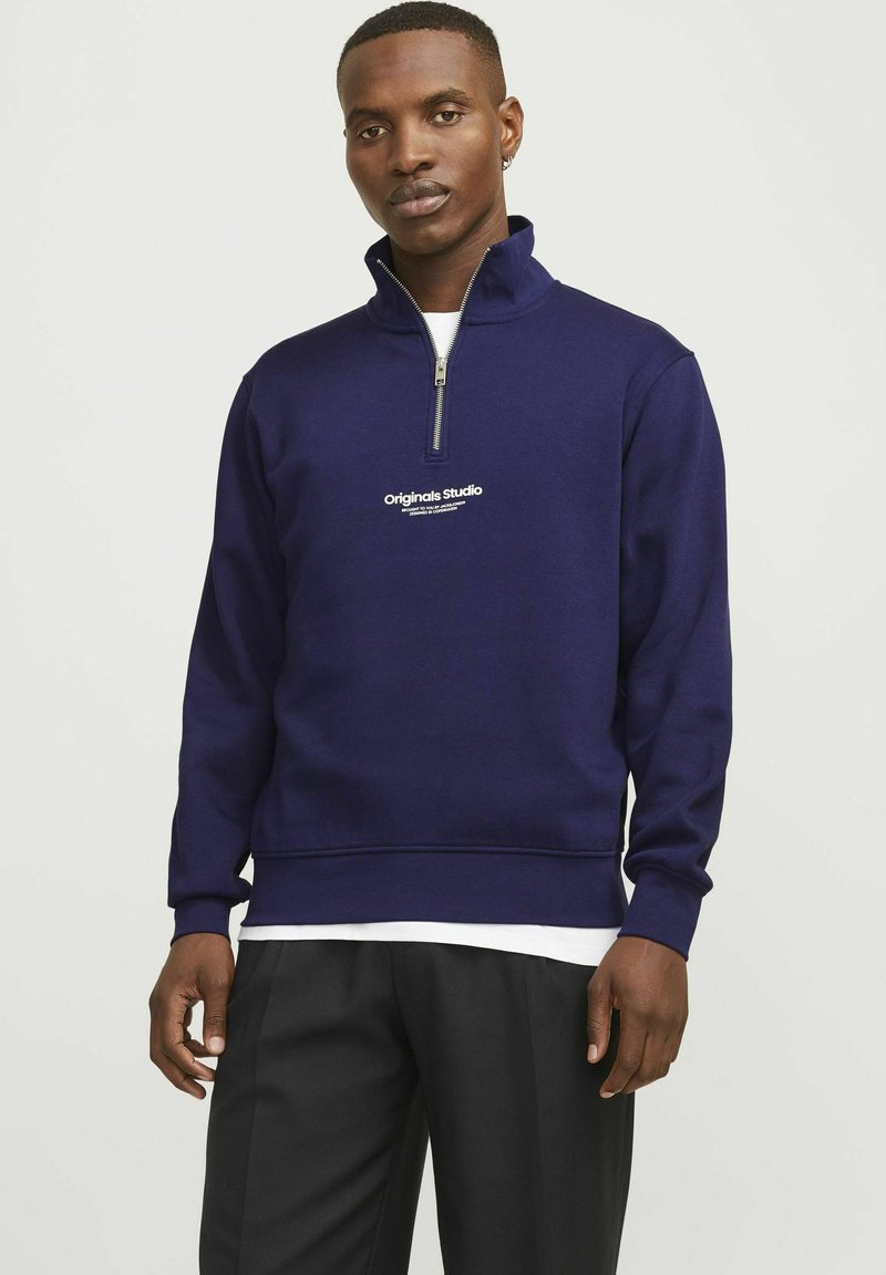 Sudadera con cremallera azul marino hecha de tela suave, con un alto cuello, puños acanalados y un logo impreso en blanco en el pecho.