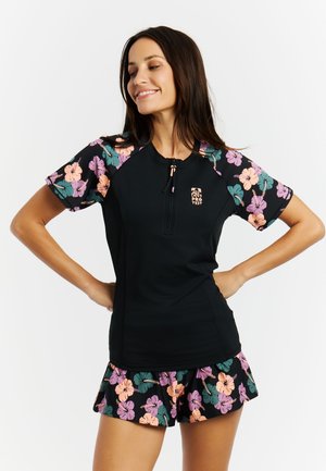 Vrouw die glimlacht met handen in de zij, gekleed in een zwarte rash guard met korte mouwen en bijpassende korte broek met bloemenprint.