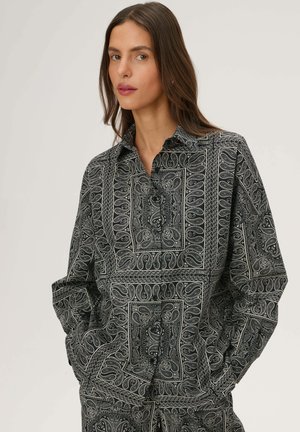 Kobieta o długich brązowych włosach, ubrana w oversize'ową koszulę w czarno-białe wzory paisley, z rękami w kieszeniach na jednolitym jasnym tle.