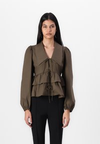 Blouse en coton marron avec des manches bouffantes, col en V et détail de nouage à l'avant. Présente des volants le long du buste et une taille cintrée.