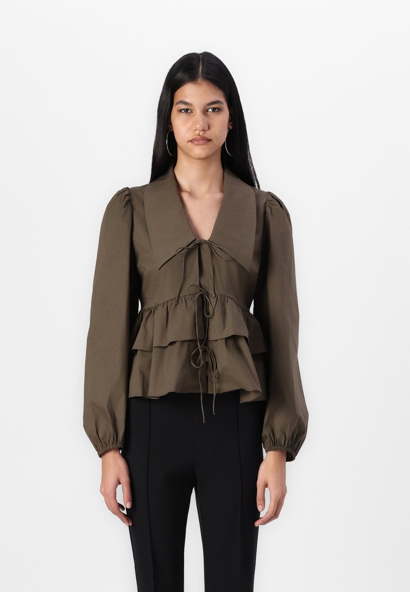 Blouse en coton marron avec des manches bouffantes, col en V et détail de nouage à l'avant. Présente des volants le long du buste et une taille cintrée.