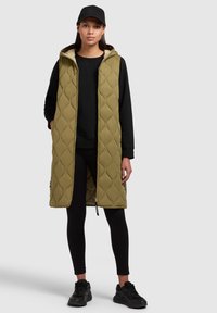 Gilet lungo verde trapuntato con cappuccio e dettagli elastici ai lati. Indossato sopra una felpa nera e leggings, abbinato a sneakers nere.