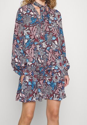 Femme portant une robe à manches longues avec un motif floral abstrait et coloré en bleu, bordeaux, blanc et noir, avec un détail de nœud au cou.