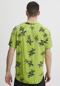 Camisa verde de manga corta con patrones florales negros, con un corte relajado y un dobladillo recto, hecha de un tejido ligero.