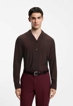 Giovane uomo con capelli corti e scuri che indossa una camicia scura a maniche lunghe con fantasia e pantaloni bordeaux con cintura nera.