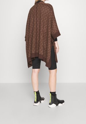 Persona con poncho marrón con estampado, pantalones cortos de ciclismo negros y zapatillas negras con suelas blancas y tiradores de neón amarillos, de pie frente a un fondo liso.