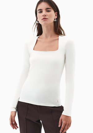 Mujer con cabello largo castaño que lleva pendientes de plata, una blusa blanca de manga larga y escote cuadrado, y pantalones marrón oscuro, de pie frente a un fondo blanco.