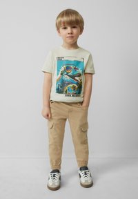 Lichtbeige T-shirt met een kleurrijke dinosaurusafbeelding. Gecombineerd met kaki cargo shorts en witte sneakers met zwarte accenten.