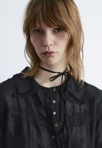 Black blouse with lace detailing, fold-down collar, and button front. Features a tied ribbon accent at the neck and a soft, textured fabric.