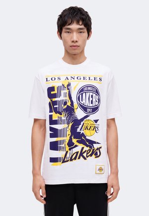 Ung mand iført en hvid Los Angeles Lakers T-shirt med basketballspiller grafik og holdlogoer i lilla og gul.