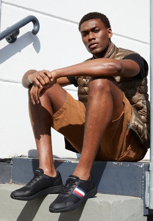 Junger Mann in braunen Shorts, schwarzem T-Shirt und ärmelloser Steppweste sitzt auf Betontreppen und trägt schwarze Sneakers mit roten, weißen und blauen Streifen.