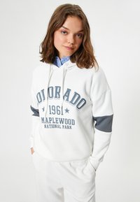 Witte hoodie van zacht materiaal met een grijze streep op de mouwen, bedrukt met de tekst "COLORADO 1961 MAPLEWOOD NATIONAL PARK" in donkergrijs.