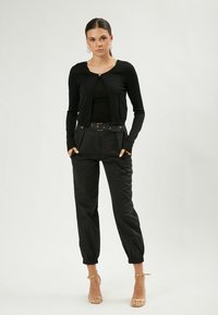 Cardigan nero a maniche lunghe con scollo rotondo e chiusura a bottoni, abbinato a pantaloni neri a vita alta con vita con cintura.