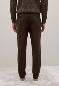 Bruine op maat gemaakte broek met een gladde textuur, met achterzakken met zwarte knoopsluiting. Het model draagt een donkere gebreide trui.