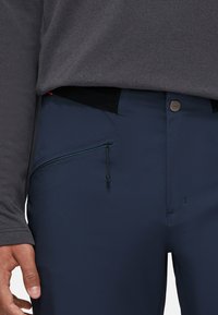 Pantalones navy con un bolsillo con cremallera, hechos de material ligero. Presentan un cierre de botón y una textura suave y mate. Llevados con una camisa oscura.